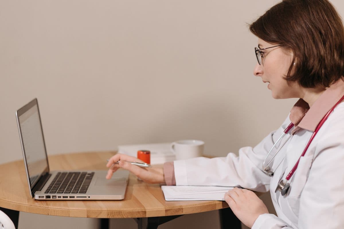 A doctor in a white coat and stethoscope reading from an open laptop at a desk.